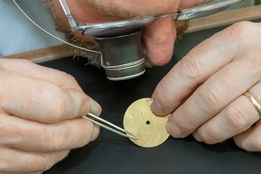 A dial blank held in tweezers under a loupe